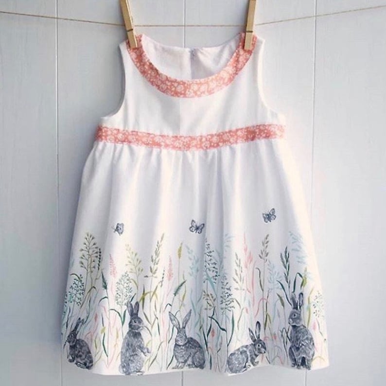 May include: A white sleeveless dress with a pink trim and a floral print of bunnies and butterflies. The dress has a gathered skirt and a round neckline.