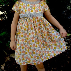 May include: A white dress with a floral print in shades of yellow, orange, and pink. The dress has a gray polka dot trim around the neckline and a gray ribbon tied at the waist.