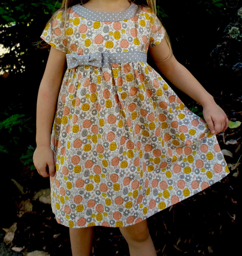 May include: A white dress with a floral print in shades of yellow, orange, and pink. The dress has a gray polka dot trim around the neckline and a gray ribbon tied at the waist.