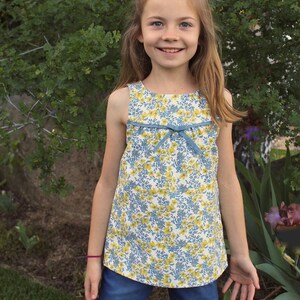 May include: A young girl wearing a sleeveless floral dress with a blue bow and denim shorts. She is standing in front of a green bush.