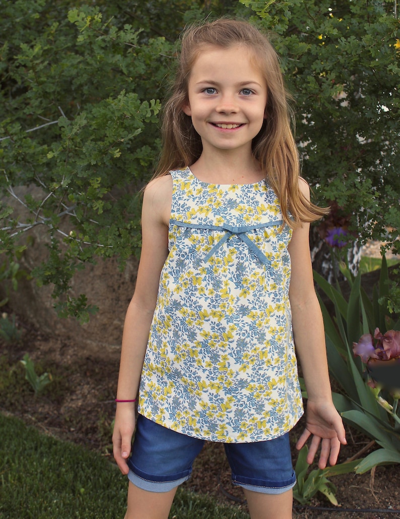 May include: A young girl wearing a sleeveless floral dress with a blue bow and denim shorts. She is standing in front of a green bush.