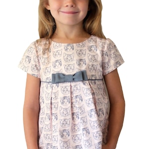 May include: A pink and white short-sleeved dress with a gray bow and a pattern of cat faces. The dress has a pleated skirt and a pocket on the left side.
