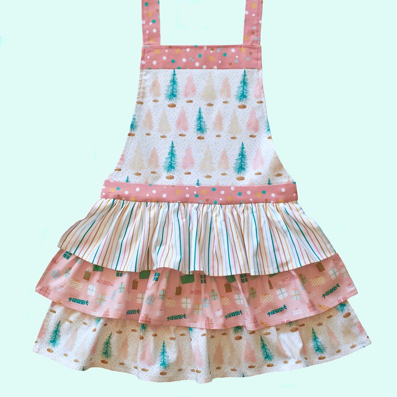May include: A pink and white tiered apron with a Christmas tree print. The apron has a ruffled bottom and a pink and white polka dot trim.
