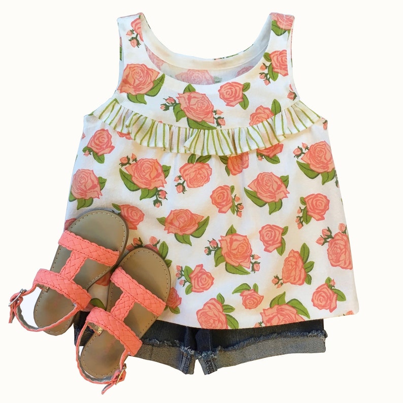 May include: A white tank top with a pink floral print and a green ruffle trim. The tank top is paired with denim shorts and pink sandals.