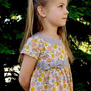 May include: A young girl wearing a white dress with a floral print and a gray bow. The dress has a gray and white polka dot trim around the neckline.