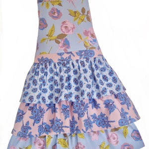 May include: A blue and pink floral print apron with a ruffled skirt. The apron has a pink top section and a blue bottom section with three tiers of ruffles.