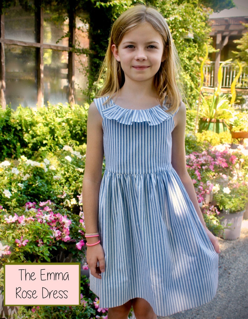 May include: A blue and white striped dress with a ruffled neckline. The dress is sleeveless and has a flared skirt. The text "The Emma Rose Dress" is in a pink box in the lower left corner. The girl is wearing a pink bracelet.