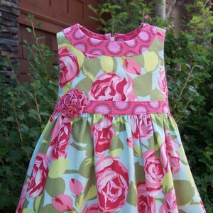 May include: A sleeveless dress with a pink floral print on a light blue background. The dress has a pink collar and a pink ribbon with a flower accent at the waist.