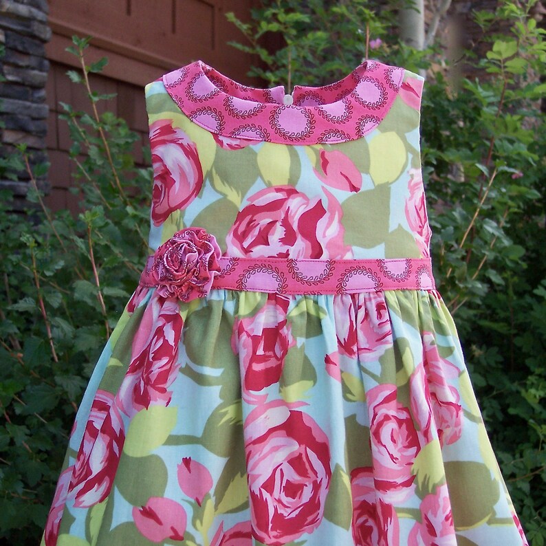 May include: A sleeveless dress with a pink floral print on a light blue background. The dress has a pink collar and a pink ribbon with a flower accent at the waist.