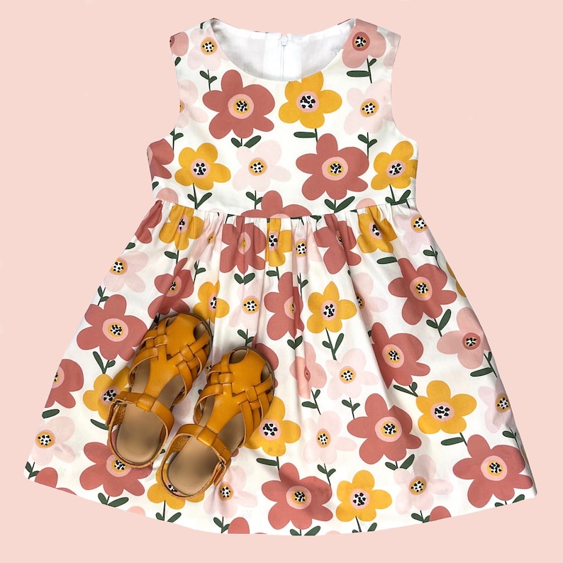 Puede incluir: Un vestido blanco sin mangas con estampado floral de flores rosas y amarillas. El vestido tiene una falda fruncida y se combina con dos sandalias de cuero marr&oacute;n.
