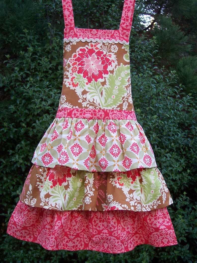 May include: A tiered apron with a brown and pink floral print. The apron has a ruffled bottom and a brown ribbon around the waist.