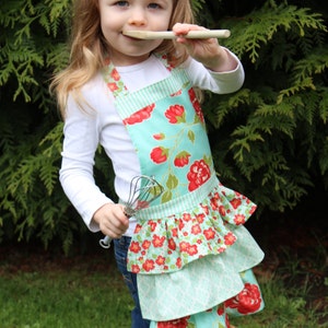 May include: A young child wearing a blue and white floral apron with a ruffled bottom. The apron has a white and blue striped waistband. The child is holding a wooden spoon and a whisk.