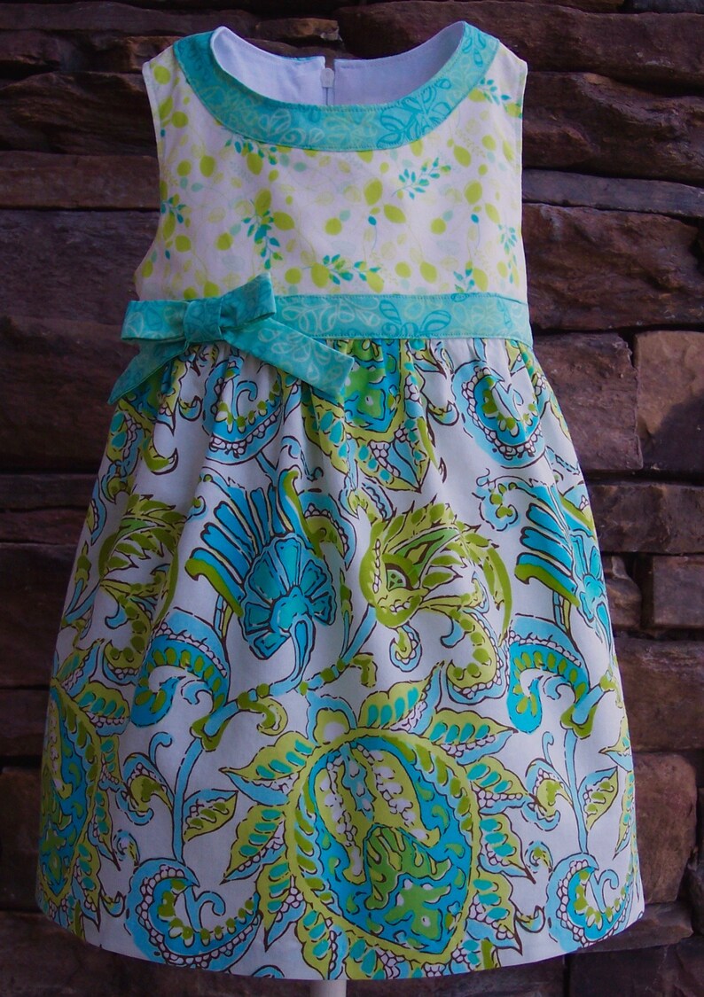 May include: A white dress with a green and blue floral pattern. The dress has a green and blue ribbon tied at the waist.