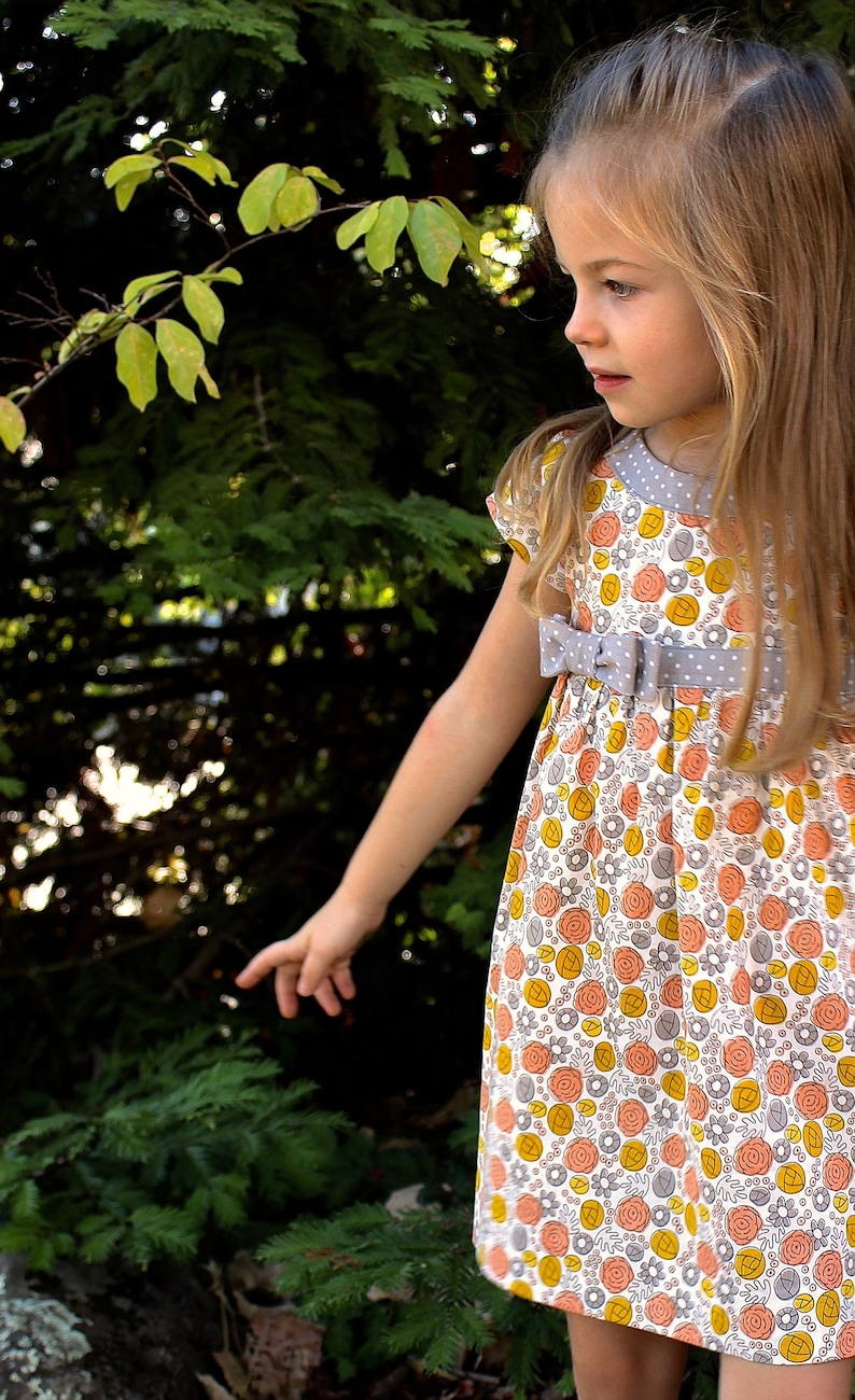 May include: A young girl wearing a white dress with a floral pattern in shades of orange, yellow, and gray. The dress has a gray ribbon tied at the waist and a white polka dot pattern on the ribbon. The girl is standing in front of a green bush.