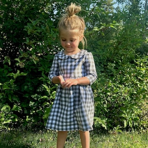 Puede incluir: Una ni&ntilde;a peque&ntilde;a con un vestido de cuadros grises y blancos con un cuello blanco de Peter Pan. El vestido tiene una falda fruncida y mangas 3/4.