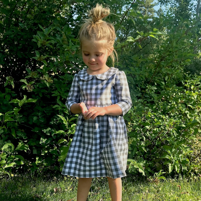 Puede incluir: Una ni&ntilde;a peque&ntilde;a con un vestido de cuadros grises y blancos con un cuello blanco de Peter Pan. El vestido tiene una falda fruncida y mangas 3/4.