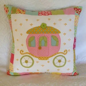 Könnte beinhalten: Ein weißes Kissen mit einem bunten Patchwork-Design, das eine rosafarbene und gelbe Kutsche mit grünen Akzenten zeigt. Das Kissen hat einen weißen Hintergrund mit einem verstreuten Muster aus kleinen gelben Blumen.