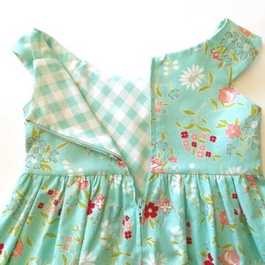 May include: A close-up of a turquoise dress with a floral pattern in pink, red, and white. The dress has a white and light blue gingham lining and a zipper down the back. The dress has short sleeves.