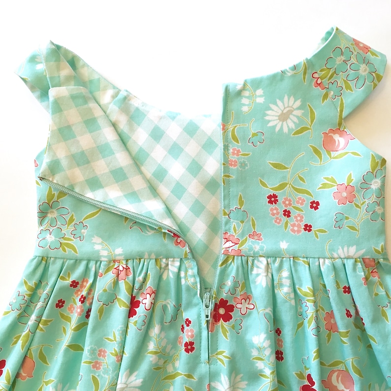 May include: A close-up of a turquoise dress with a floral pattern in pink, red, and white. The dress has a white and light blue gingham lining and a zipper down the back. The dress has short sleeves.