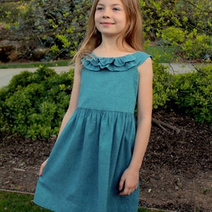 May include: A teal-colored sleeveless dress with a ruffled neckline. The dress has a gathered waist and a flared skirt. The girl is standing outside in front of green foliage.