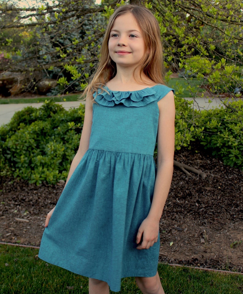 May include: A teal-colored sleeveless dress with a ruffled neckline. The dress has a gathered waist and a flared skirt. The girl is standing outside in front of green foliage.