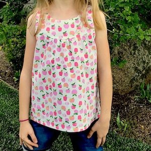 May include: A sleeveless white tank top with a pink and white apple print. The tank top has a gathered neckline and a relaxed fit.