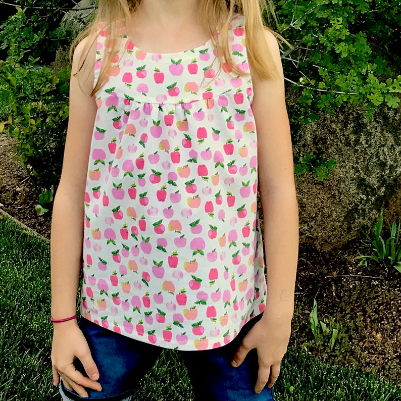 May include: A sleeveless white tank top with a pink and white apple print. The tank top has a gathered neckline and a relaxed fit.