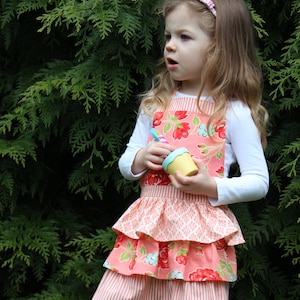 May include: A young girl wearing a pink and white floral print apron with a ruffled bottom. She is holding a small yellow pot in her hands.