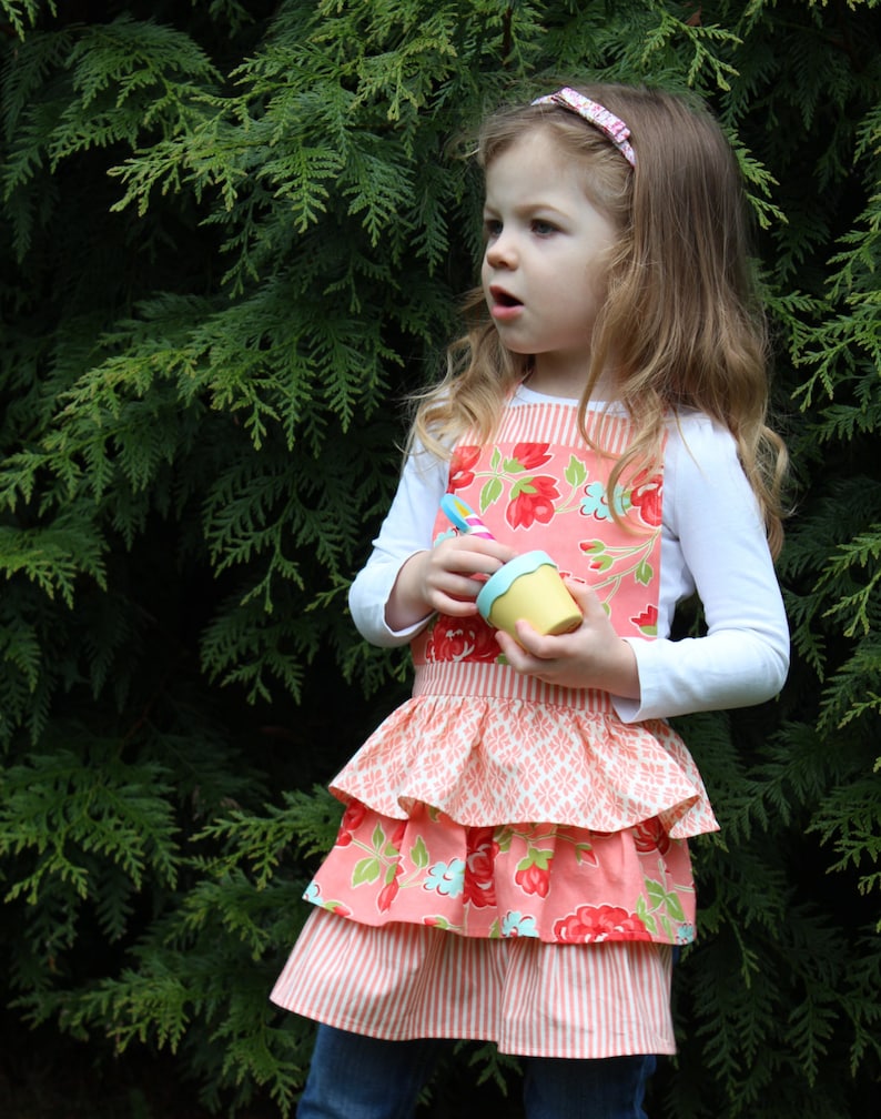 May include: A young girl wearing a pink and white floral print apron with a ruffled bottom. She is holding a small yellow pot in her hands.