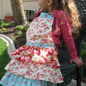 May include: A young girl wearing a multi-layered floral print apron with a ruffled hem. The apron has a blue and white top layer, a red and white middle layer, and a white and green bottom layer. The girl is wearing black leggings and a pink long-sleeved shirt.