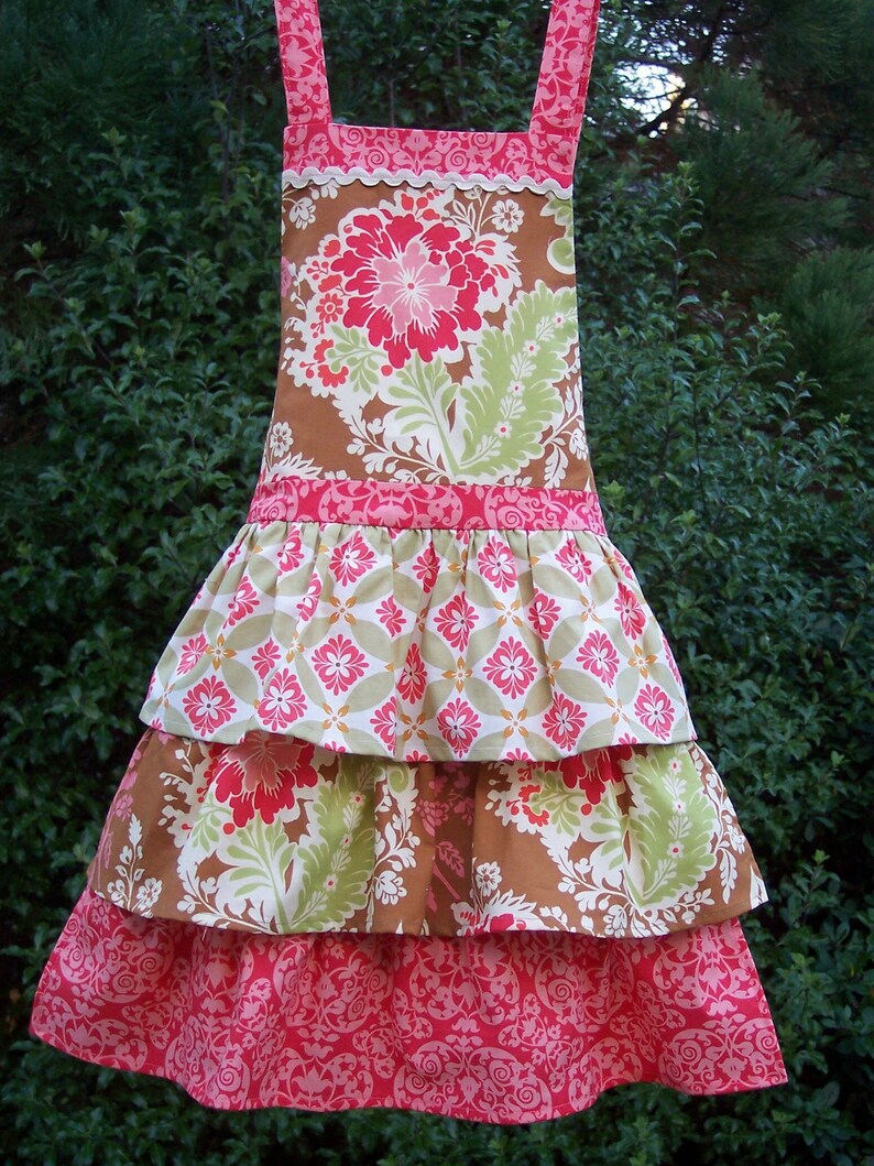 May include: A tiered apron with a brown, green, and pink floral print. The apron has a ruffled bottom and a scalloped top edge.