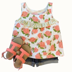 May include: A white tank top with a pink floral print and a green ruffle trim. The tank top is paired with denim shorts and pink sandals.
