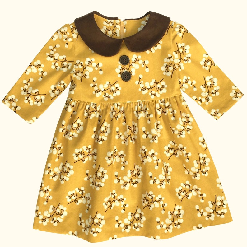Puede incluir: Un vestido amarillo con estampado floral blanco y cuello de pana marr&oacute;n. El vestido tiene mangas largas y cierre de bot&oacute;n en el cuello.