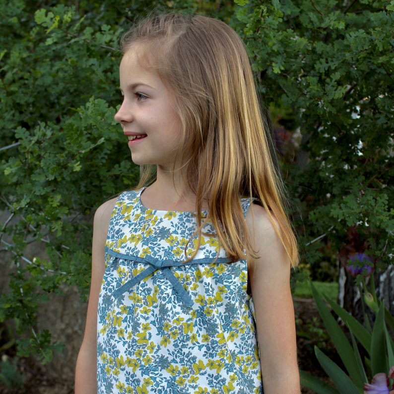 May include: A young girl with long blonde hair is wearing a sleeveless dress with a blue and yellow floral pattern. The dress has a blue ribbon tied at the waist.