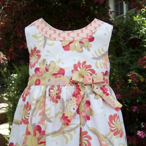 May include: A white sleeveless dress with a floral print in shades of pink, yellow, and gray. The dress has a bow at the waist and a contrasting trim around the neckline.