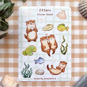 Könnte beinhalten: Ein Stickerbogen mit Cartoon-Ottern, Fischen, Seetang und Muscheln. Der Text auf dem Bogen lautet "Otters Sticker Sheet" und "made by exozetart". Der Hintergrund ist ein hellgrünes Gittermuster. Die Otter sind in verschiedenen Posen.