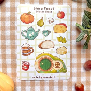 May include: A sticker sheet with the text "Shire Feast" features illustrations of food items like a pumpkin, apple, pear, mushroom, cheese, bread, and a teapot. A hobbit hole and a jar of jam are also depicted. The sticker sheet is labeled "made by exozetart".