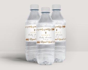 Op de afbeelding: Drie doorzichtige plastic waterflessen met witte doppen. Elke fles heeft een etiket met de tekst "Nora's baby shower 2.20.23" en een bijengrafiek, omringd door gouden en bruine bloemen- en bladeraccenten.