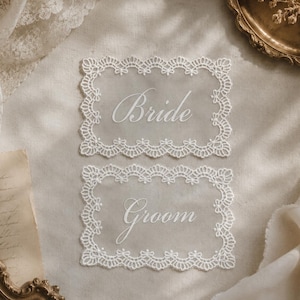 May include: Two rectangular white lace signs with scalloped edges. The top sign features the word Bride in a white cursive script, and the bottom sign features the word Groom in a white cursive script, both centred on a sheer white lace background.