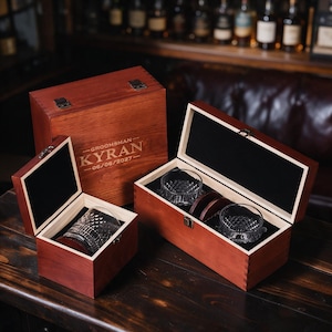 Peut inclure: Trois boîtes en bois foncé avec couvercles à charnières et doublure intérieure noire. Une grande boîte est gravée GROOMSMAN KYRAN 06/06/2027. Deux boîtes ouvertes plus petites contiennent des verres à whisky en cristal texturé et des sous-verres.