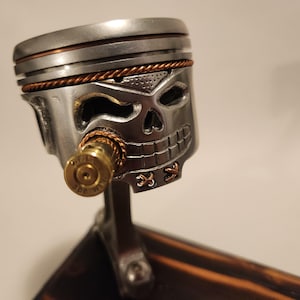 May include: A handcrafted metal sculpture of a skull, made from a repurposed engine piston. The skull features a detailed design with copper wire accents and a bullet as a decorative element. The sculpture is mounted on a dark wooden base.