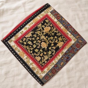 May include: A square scarf featuring a central black panel with a gold and green floral pattern, surrounded by a red border, a cream-colored band with gold floral motifs, a black outer border, and a grey edge with red and gold floral accents.