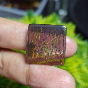 May include: A square-shaped boulder opal gemstone featuring a dark brown ironstone matrix with vibrant, vertical flashes of iridescent green, blue, and gold colour play across the polished, smooth surface of the stone.