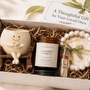 May include: A white gift box containing a cream-colored ceramic planter shaped like a smiling character on a wooden bench, a brown glass jar of Lavender Vanilla soy wax candle, a small glass bottle of matches, and a white beaded decorative ring.