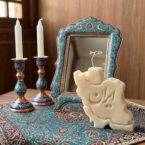 May include: A decorative arrangement featuring a cream-colored candle in the shape of a country, a framed mirror, and two candle holders with white candles. The candle holders have blue and copper accents, and the scene is set on a patterned textile.