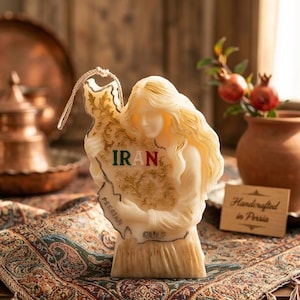 May include: A cream-colored candle shaped like a woman holding a map of Iran. The word "IRAN" is in green. The map has the words "Persian Gulf" on it. The candle is on a patterned fabric.