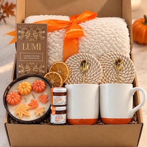 May include: A gift box containing a white sherpa blanket with an orange ribbon, a Lumi salted caramel hot chocolate bar, two white mugs with orange bases, two gold spoons, two rope coasters, two jars of multiflora honey, and a candle with pumpkin and leaf wax.