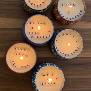 May include: Six lit candles in decorative tins with various messages spelled out in letter beads. Messages include "I AM PREGNANT", "LOVE YOU FOREVER", "YOU ARE A TRUE FIGHTER", "HAPPY ANNIVERSARY", "FIGHT HOPE BELIEVE", and "HBD SARAH".