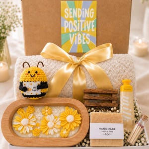 Può includere: Un set regalo contenente un'ape all'uncinetto che tiene un cartello con scritto BEE happy, una coperta testurizzata color crema con un nastro dorato, un vassoio in legno con fiori gialli, sapone artigianale, lozione al latte e fiammiferi.