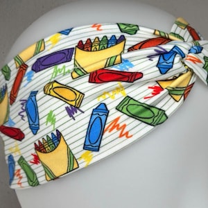 May include: White headband with a colorful crayon pattern. The crayons are scattered across the headband and some are in a box. The background is a white and green striped pattern.